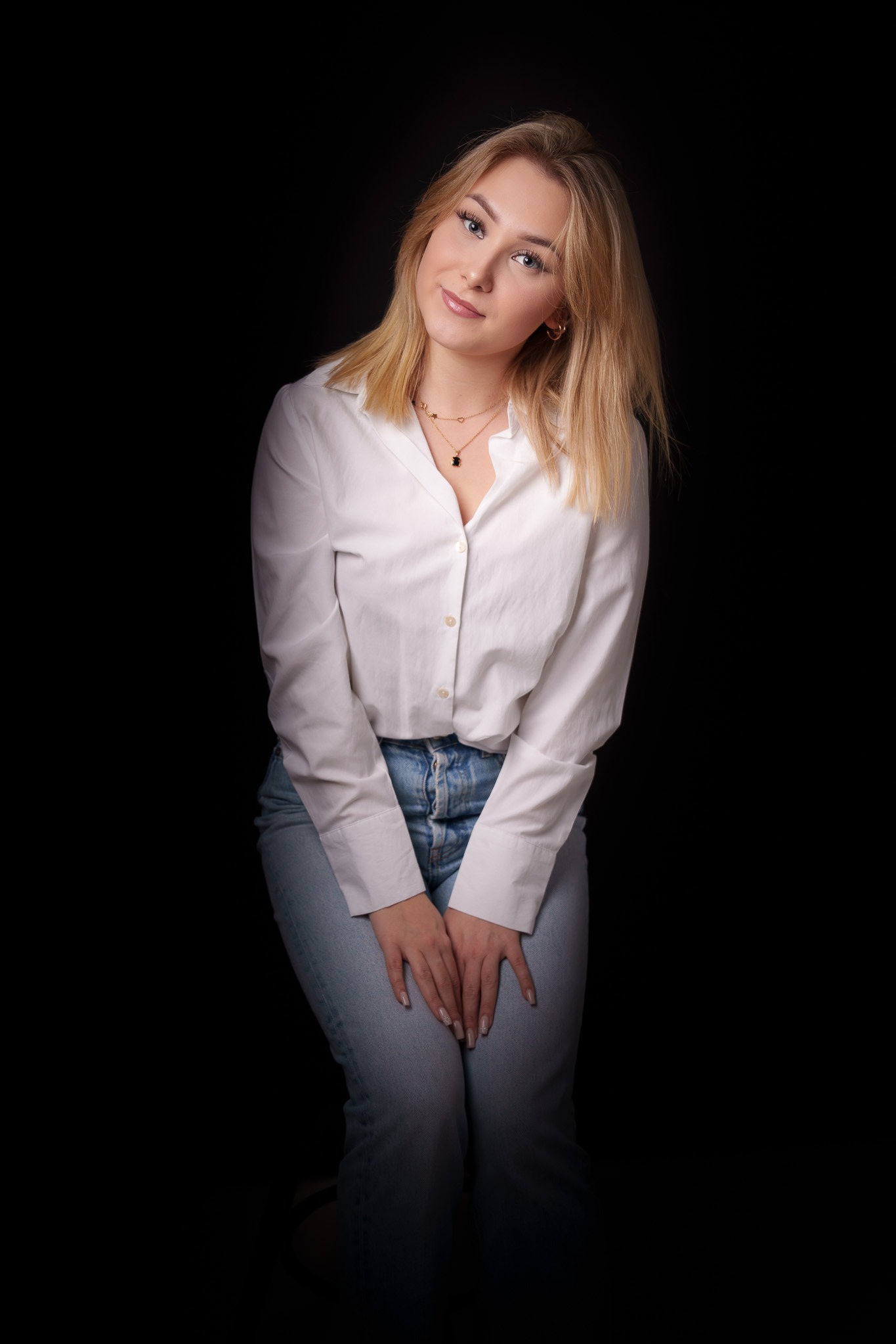 Portret młodej blondynki w białej koszuli i jeansach na czarnym tle, z delikatnym uśmiechem i złotą biżuterią, siedzącej na krześle.