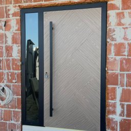 Royal Jacek Kowalczyk - Nowoczesne drzwi zewnętrzne z długim uchwytem i szybą, zamontowane w surowym murze z czerwonej cegły. Widoczne przewody i elementy montażowe.