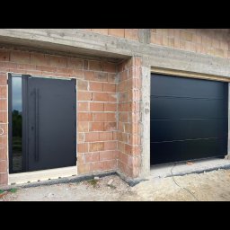 Royal Jacek Kowalczyk - Czarna brama garażowa segmentowa i drzwi zewnętrzne z przeszkleniem, zamontowane w surowym stanie budynku z czerwonej cegły. Widoczne elementy montażowe.