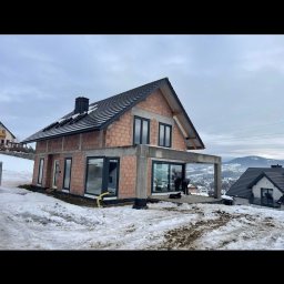 Royal Jacek Kowalczyk - Surowy stan domu z czarnymi oknami na tle zimowego krajobrazu. Widoczne cegły i betonowa konstrukcja, częściowo ośnieżone otoczenie. Prace wykończeniowe w toku.