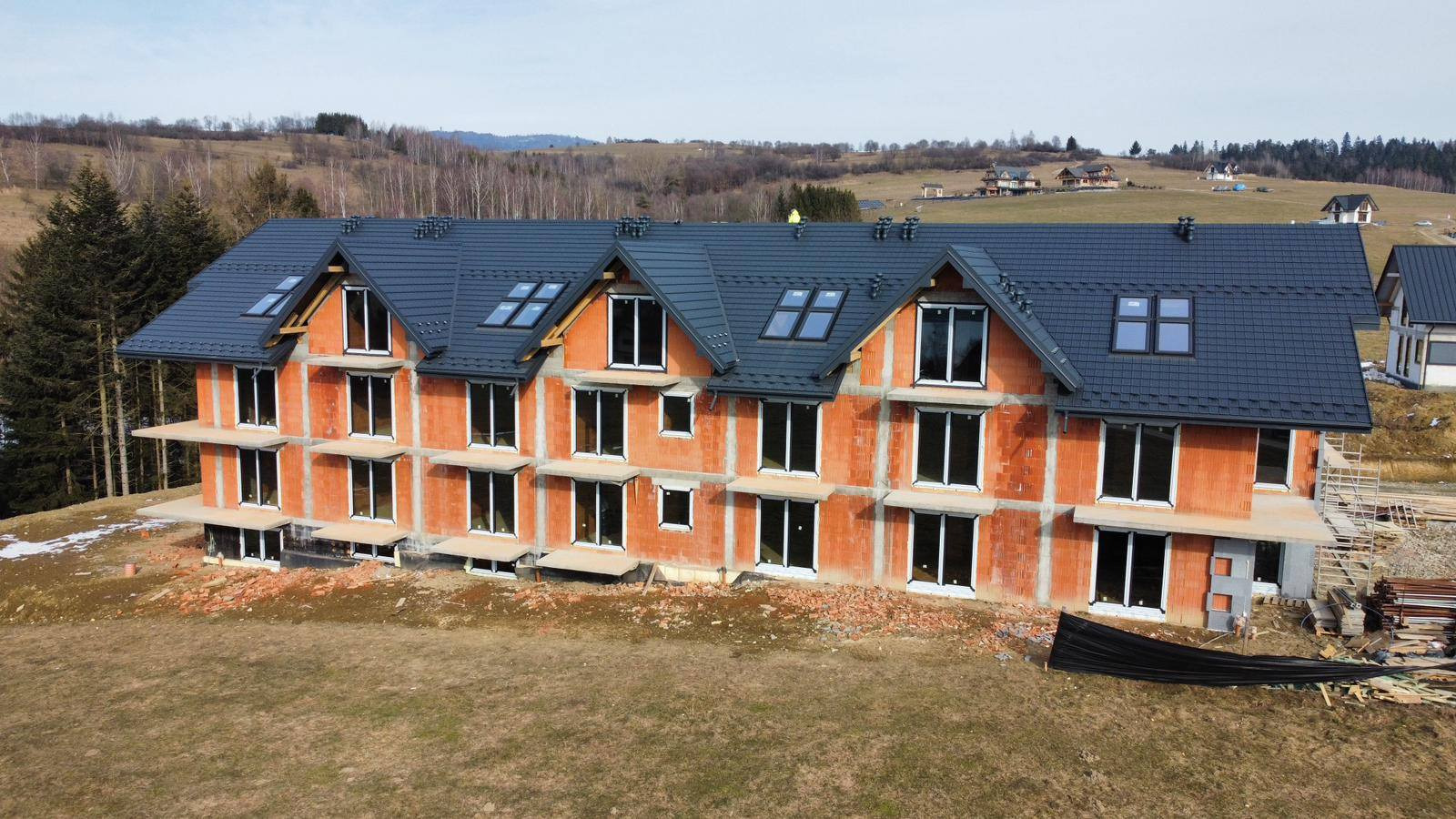 Budynek w stanie surowym otwartym z zamontowanymi oknami i dachem z dachówką, na tle pagórkowatego krajobrazu. Widoczne rusztowania i materiały budowlane.