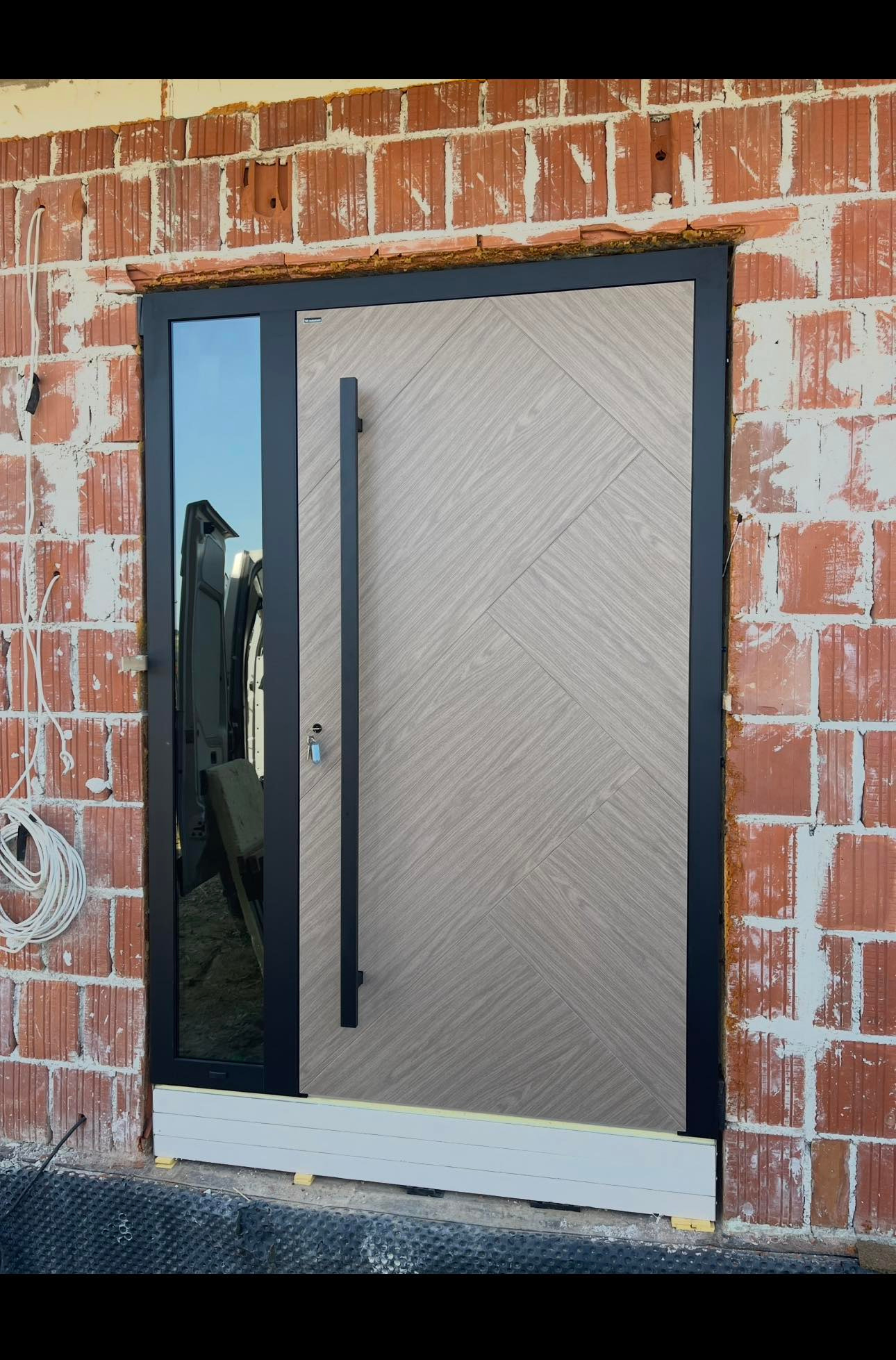 Nowoczesne drzwi zewnętrzne z długim uchwytem i szybą, zamontowane w surowym murze z czerwonej cegły. Widoczne przewody i elementy montażowe.