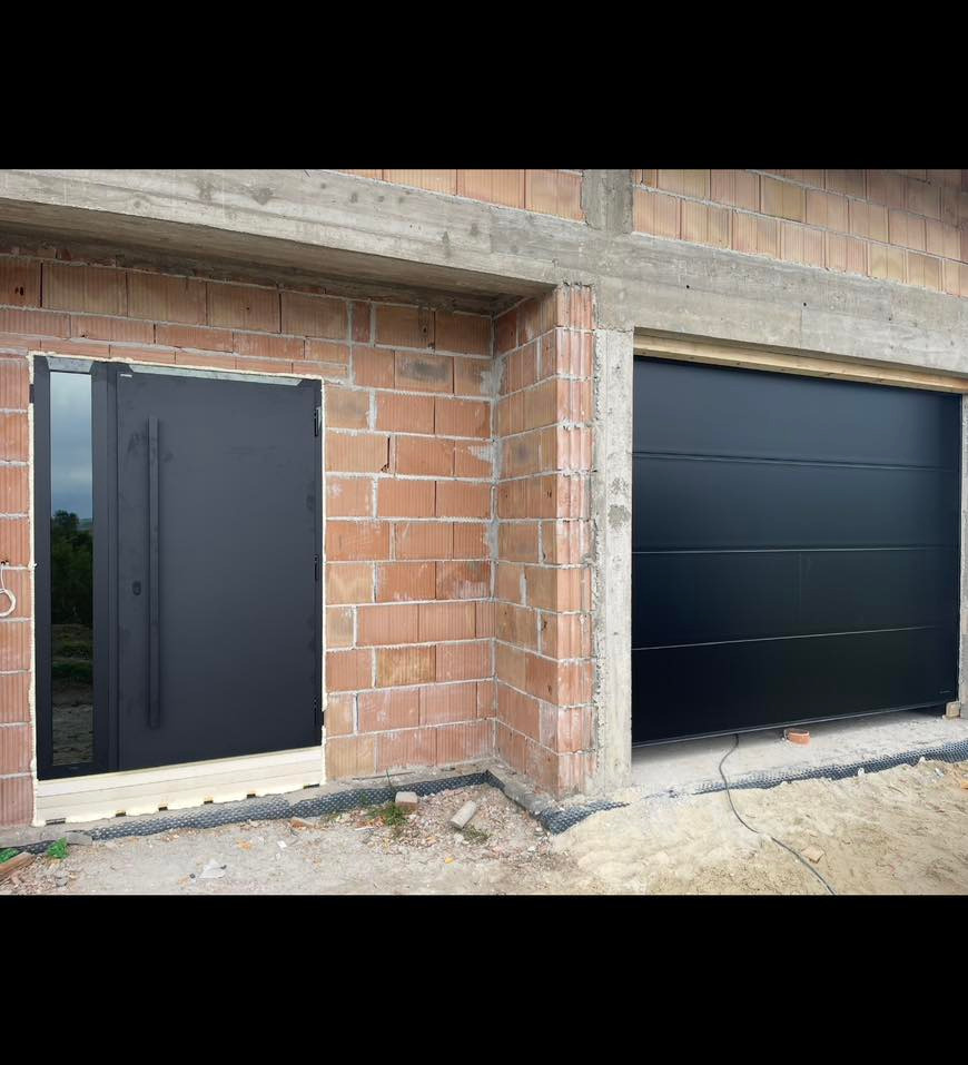 Czarna brama garażowa segmentowa i drzwi zewnętrzne z przeszkleniem, zamontowane w surowym stanie budynku z czerwonej cegły. Widoczne elementy montażowe.