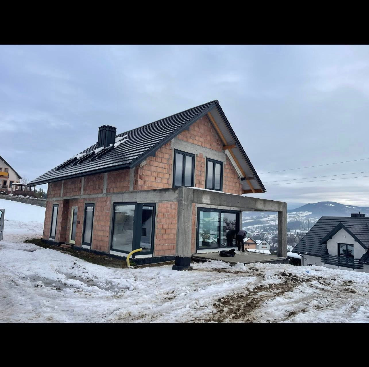 Surowy stan domu z czarnymi oknami na tle zimowego krajobrazu. Widoczne cegły i betonowa konstrukcja, częściowo ośnieżone otoczenie. Prace wykończeniowe w toku.