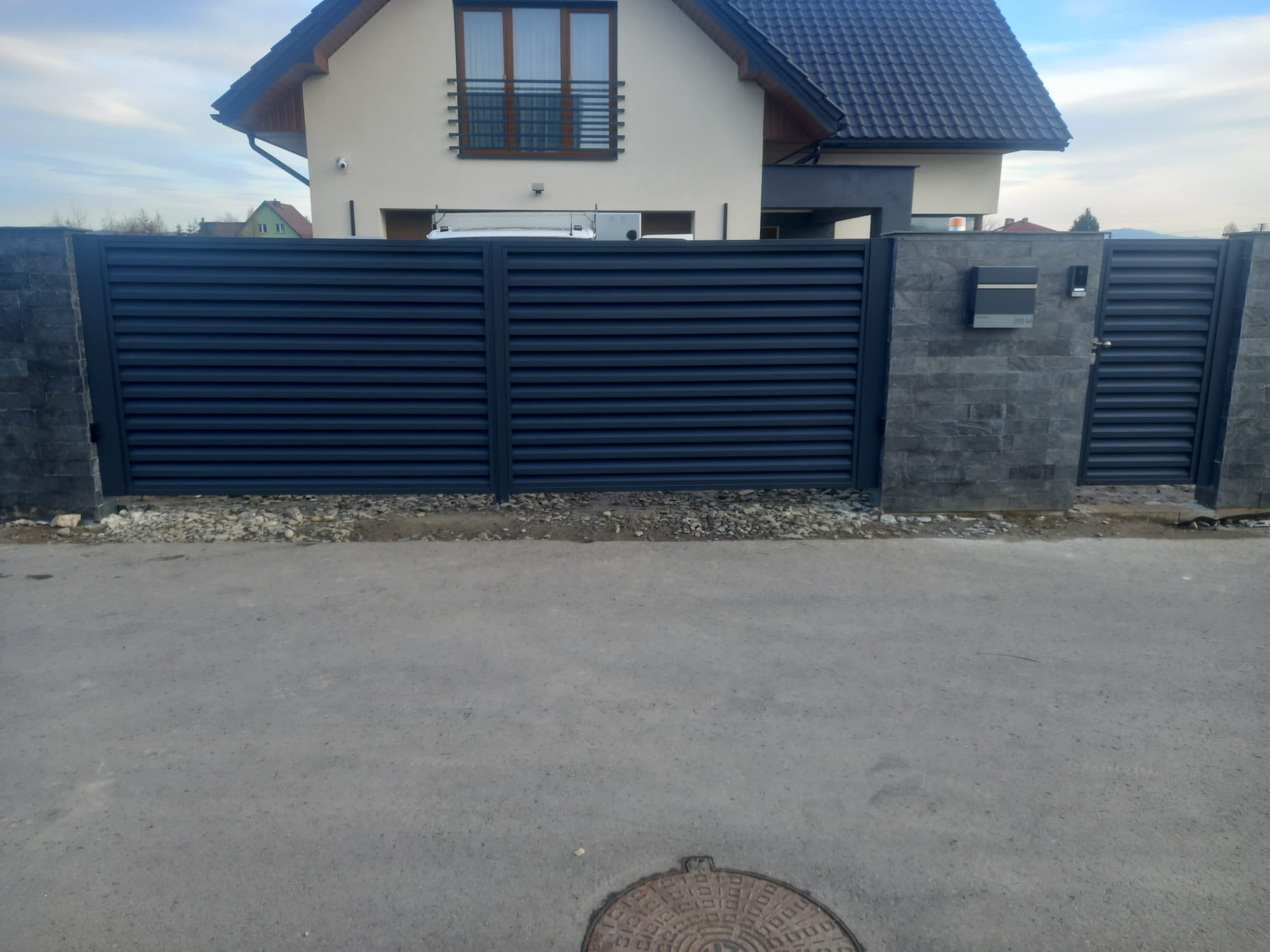 Nowoczesna, pozioma brama wjazdowa i furtka z szarego metalu, osadzone na kamiennych słupkach. W tle dom jednorodzinny z beżową elewacją i ciemnym dachem.