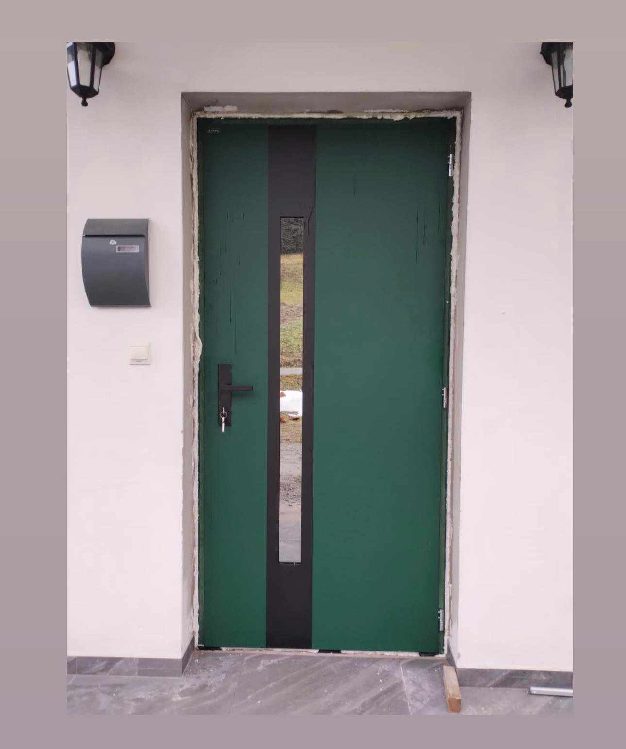 Zielone drzwi zewnętrzne z czarnym pionowym pasem i szybą, zamontowane w białej ścianie budynku w Nowym Sączu. Widoczna skrzynka na listy i lampa.