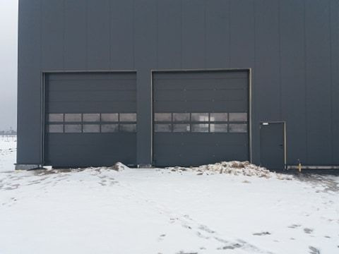Dwie szare bramy garażowe segmentowe z oknami w budynku przemysłowym, pokryte śniegiem. Obok drzwi wejściowe. Zimowy krajobraz w tle.