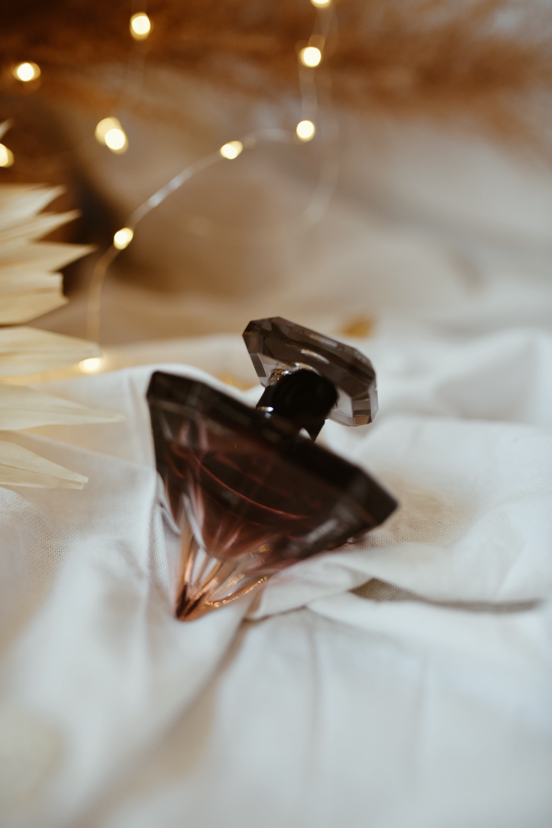 Szklany flakon perfum o geometrycznym kształcie na białym, pogniecionym materiale, z dekoracyjnymi suszonymi trawami i ciepłymi światełkami w tle.