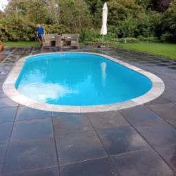 Niebieski basen ogrodowy otoczony ciemnoszarą kostką brukową, z widocznym zestawem mebli ogrodowych z rattanu i złożonym białym parasolem w tle, mężczyzna w niebieskiej koszulce przechodzi obok.