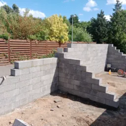 Wznoszenie stopniowanej ściany oporowej z szarych bloczk&oacute;w betonowych, widoczny fragment drewnianego ogrodzenia i narzędzia budowlane na placu budowy.