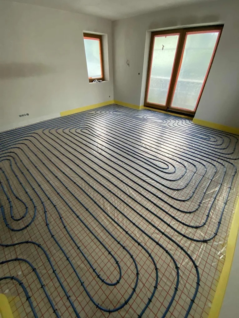 Niebieskie rury ogrzewania podłogowego ułożone na folii z czerwoną siatką w jasnym pokoju z oknami. Żółta taśma zabezpieczająca wzdłuż ścian.