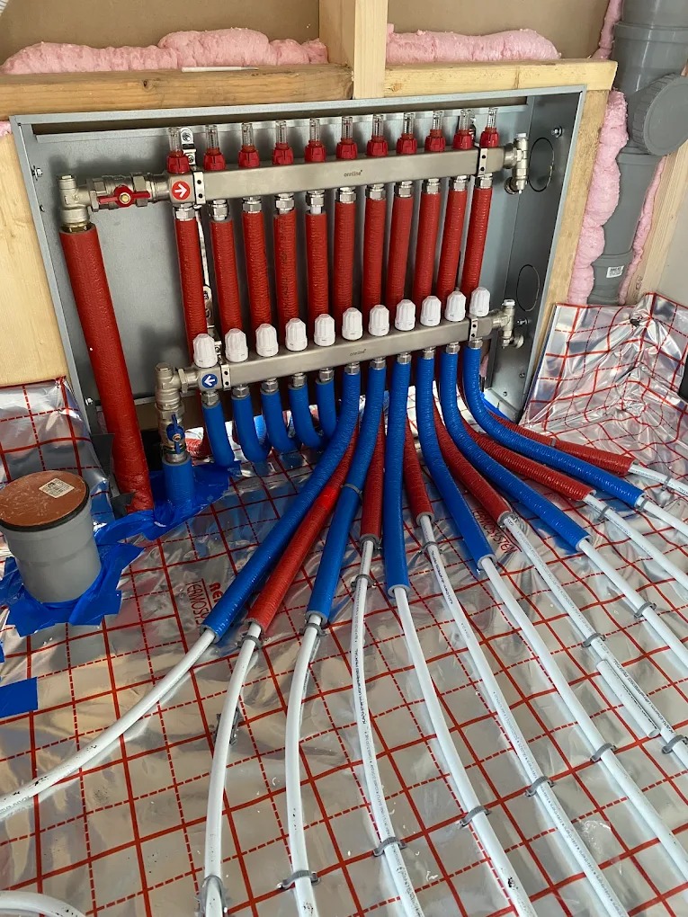 Rozdzielacz ogrzewania podłogowego z rurami w izolacji czerwonej i niebieskiej, ułożonymi na folii termoizolacyjnej z siatką. Widoczne elementy montażowe i izolacja ściany.