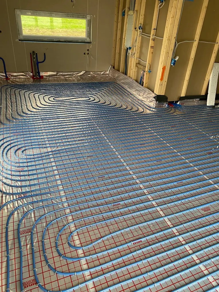 Niebieskie rury ogrzewania podłogowego ułożone na izolacji z czerwoną kratką w nowym budynku z widoczną drewnianą konstrukcją ściany i oknem w tle.