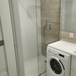 Nowoczesna łazienka z kabiną prysznicową typu walk-in, wykończoną białymi płytkami i panelami imitującymi drewno, z pralką Whirlpool umieszczoną obok kabiny.