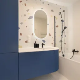 Nowoczesna łazienka z niebieską szafką pod umywalką, lustrem z podświetleniem LED, płytkami w stylu terrazzo na ścianie i czarną armaturą.