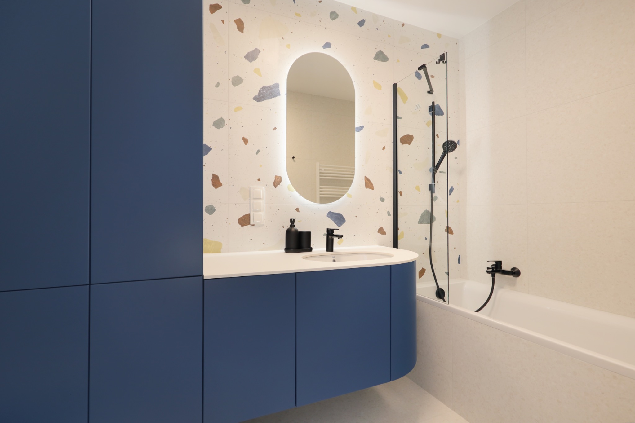Nowoczesna łazienka z niebieską szafką pod umywalką, lustrem z podświetleniem LED, płytkami w stylu terrazzo na ścianie i czarną armaturą.