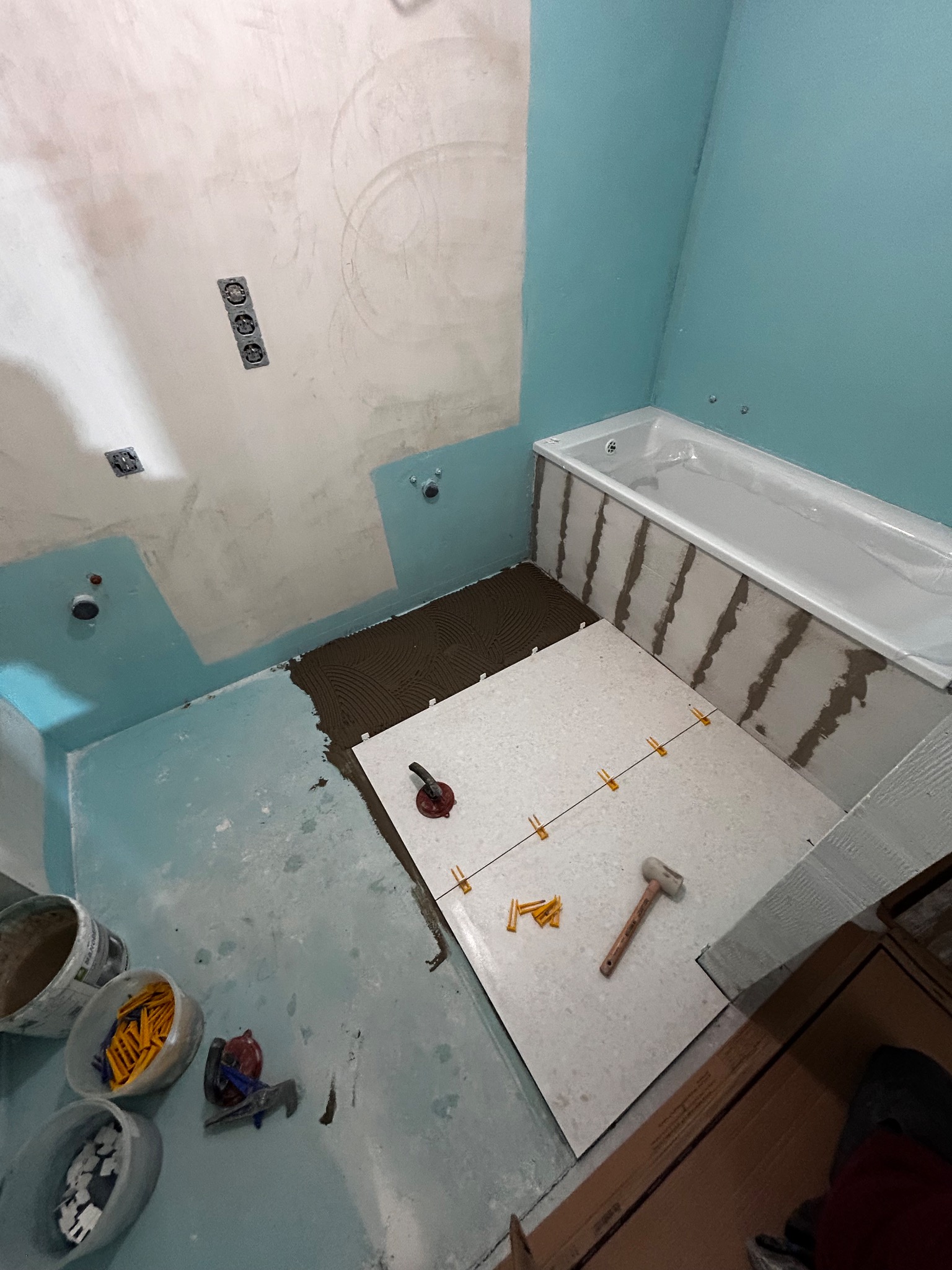 Układanie jasnych płytek podłogowych w łazience w trakcie remontu; widoczne narzędzia glazurnicze, krzyżyki dystansowe, gładzie na ścianach i przygotowana powierzchnia pod płytki.