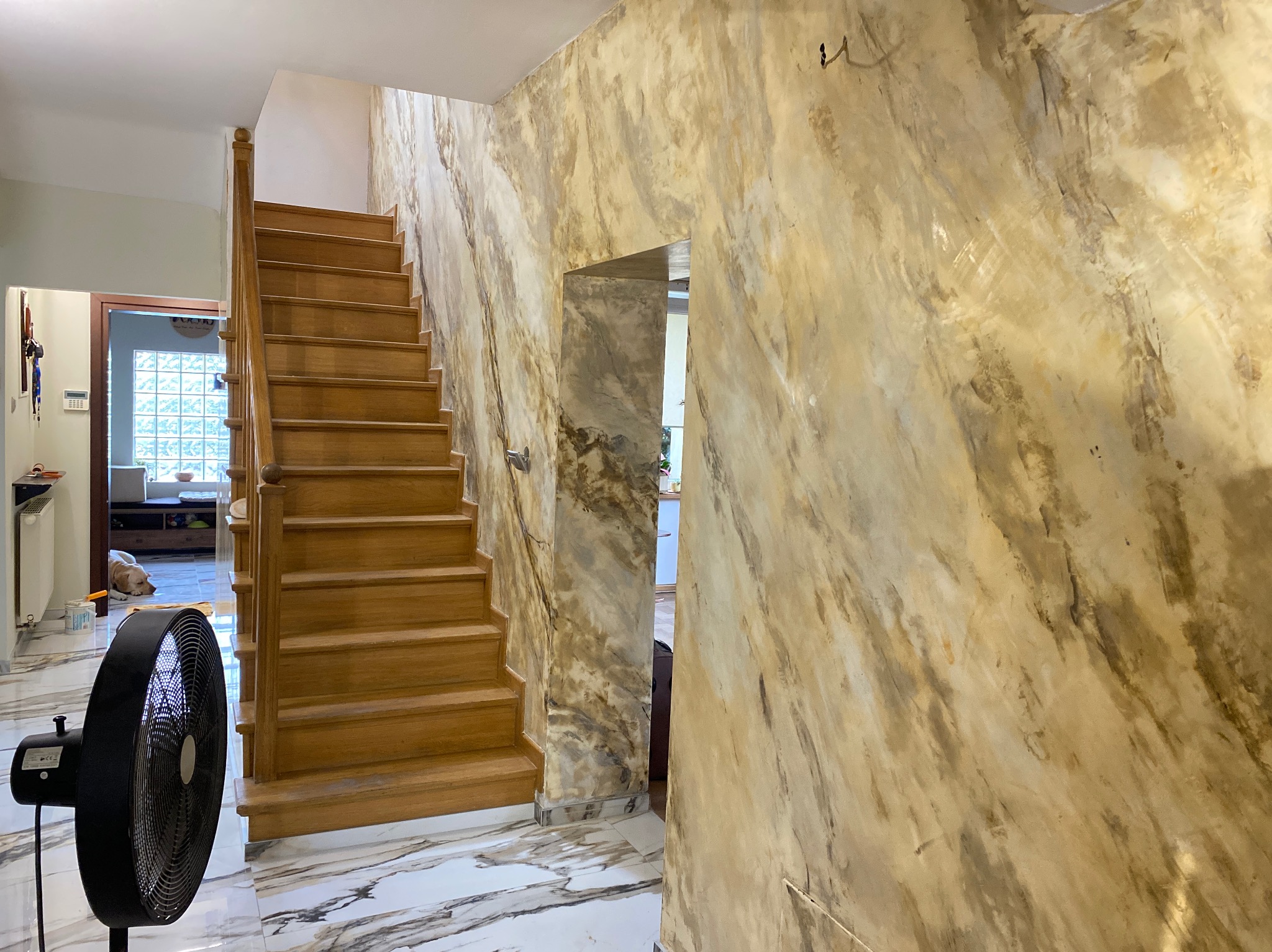 Korytarz z drewnianymi schodami i ścianami imitującymi marmur, wentylator stojący w rogu, podłoga z białego marmuru.