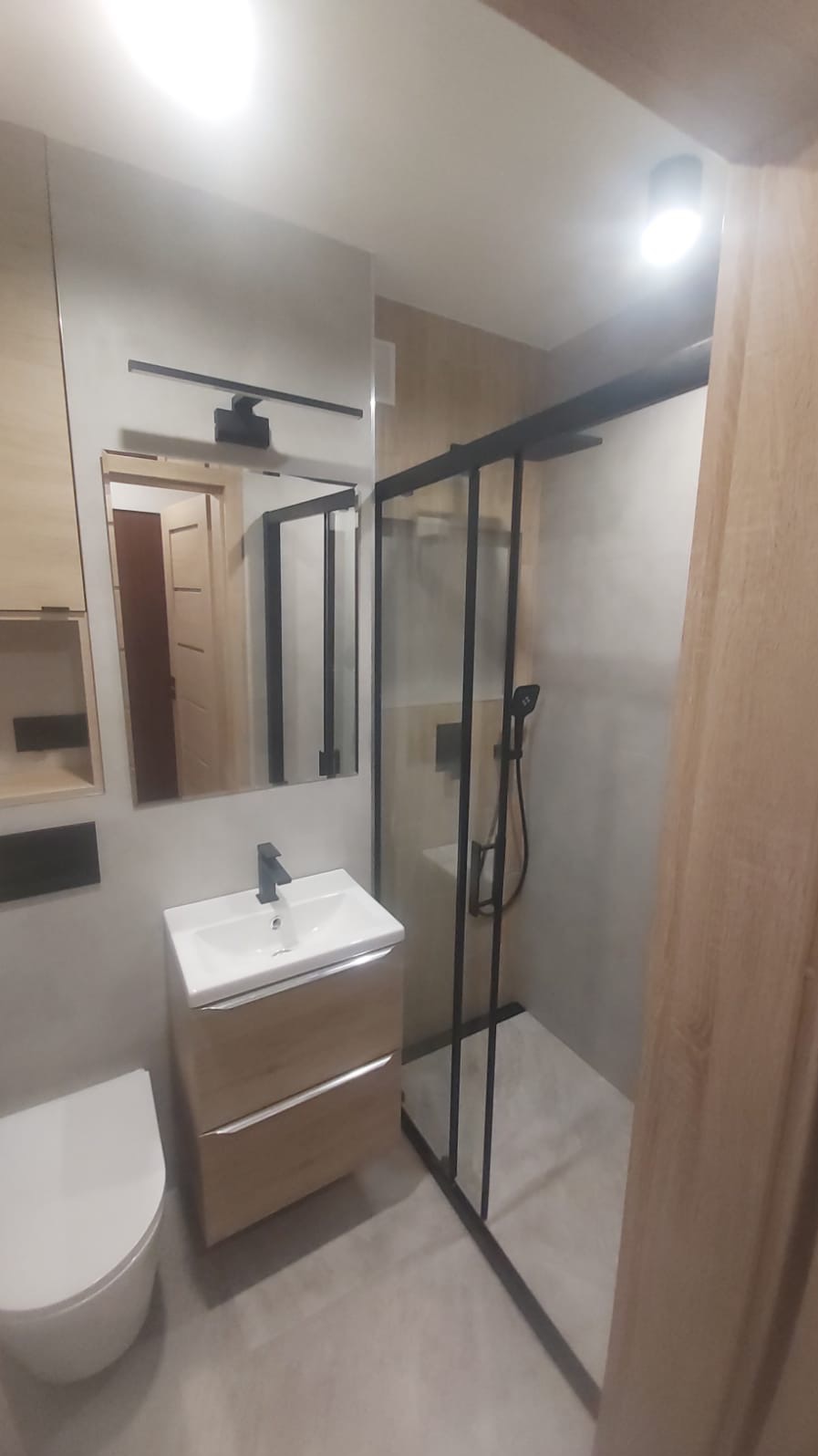 Nowoczesna łazienka z kabiną prysznicową typu walk-in z czarnymi profilami, umywalką nablatową na szafce z drewnopodobnym frontem i wiszącą miską WC, odbicie drzwi w lustrze.