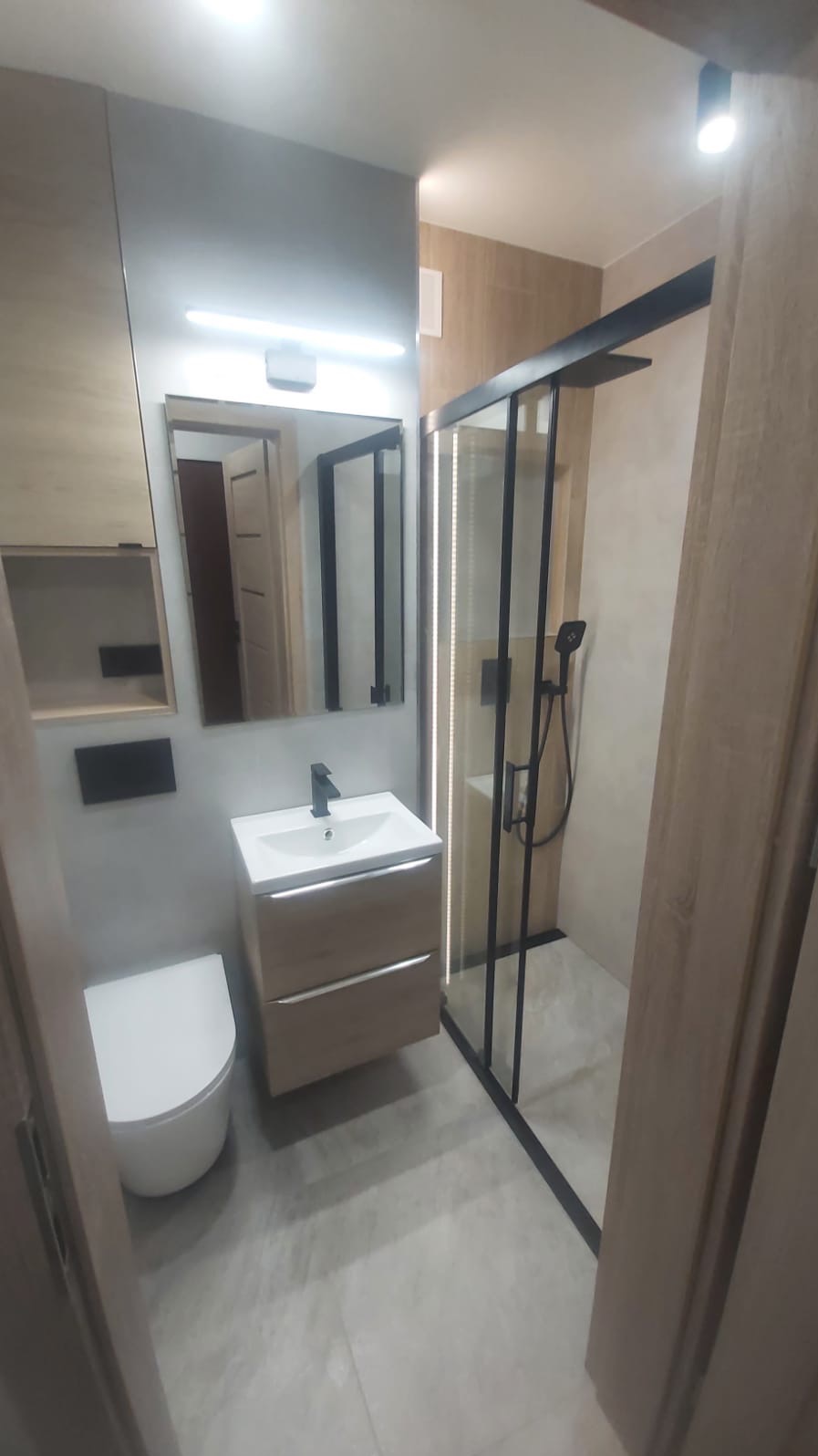 Nowoczesna łazienka z kabiną prysznicową typu walk-in z czarnymi profilami, umywalką nablatową na szafce z drewnopodobnym fornirem oraz wiszącą toaletą.