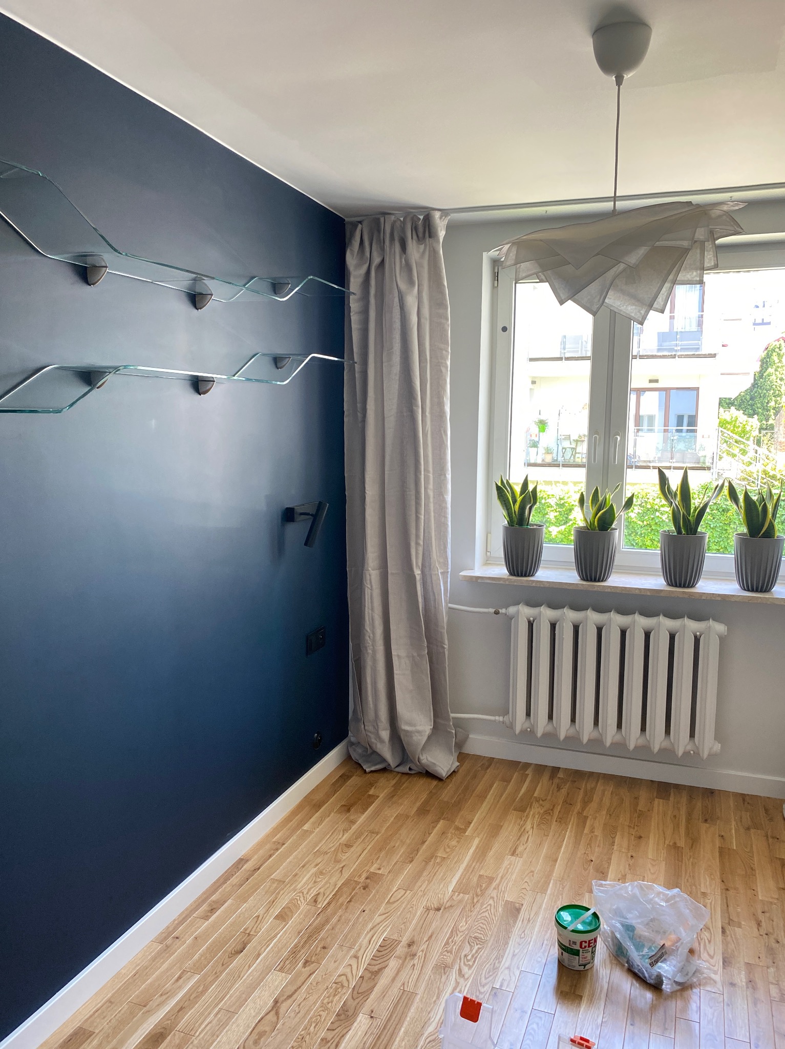 Pomieszczenie po malowaniu ściany na granatowo, z zamontowanymi szklanymi półkami, widoczne rośliny w doniczkach na parapecie, podłoga z drewnianych paneli, ślady farby i akcesoria malarskie.