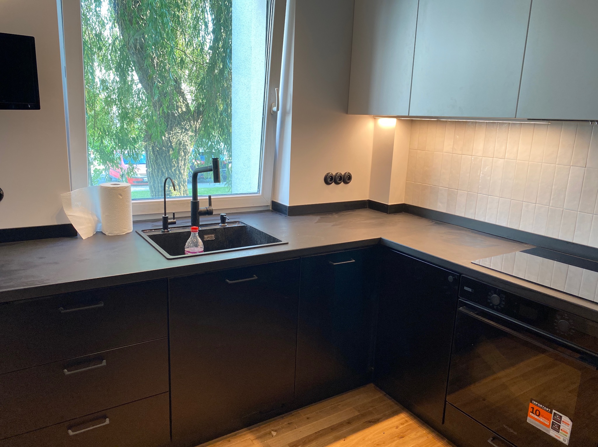 Nowoczesna, czarno-biała kuchnia z widokiem na zieleń za oknem, czarny zlew i bateria, białe płytki na ścianie, czarne szafki dolne i jasne górne, z naklejką na piekarniku.