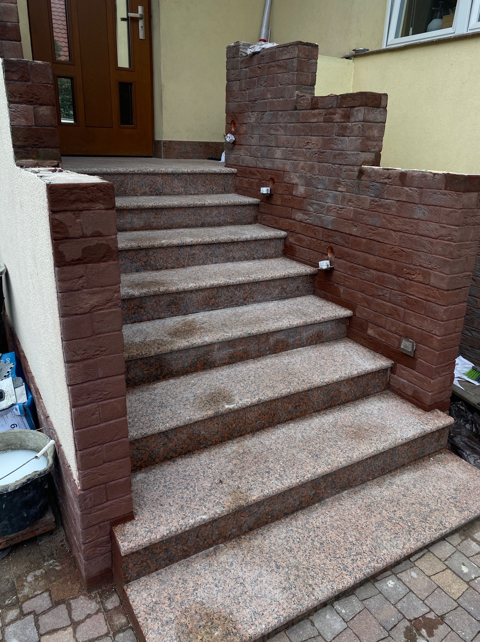 Granitowe schody wejściowe z ceglaną okładziną na ścianach bocznych, z widocznymi punktami oświetleniowymi i śladami prac wykończeniowych.