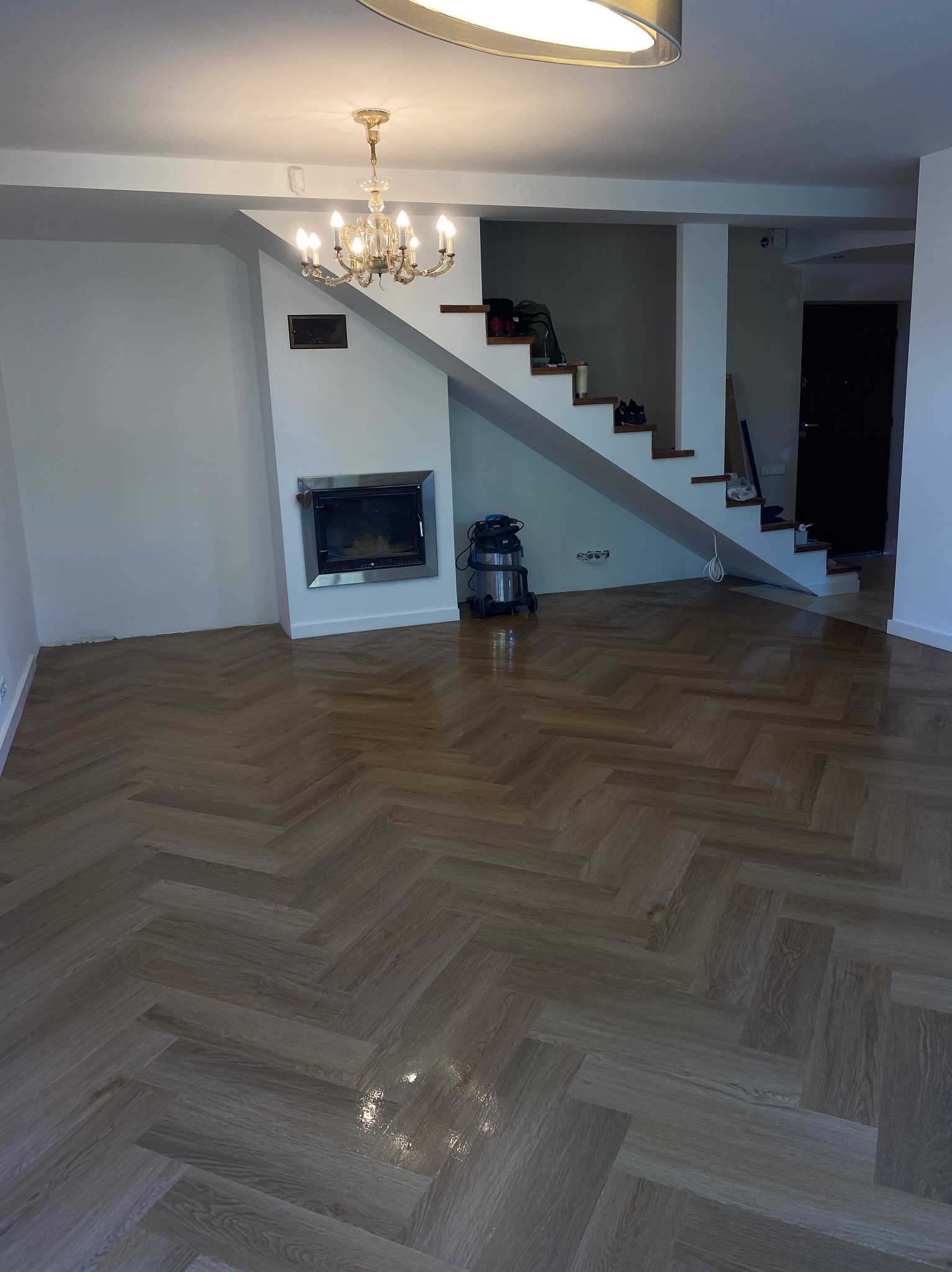 Salon po remoncie z parkietem ułożonym w jodełkę, kominkiem i schodami, widoczny odkurzacz przemysłowy