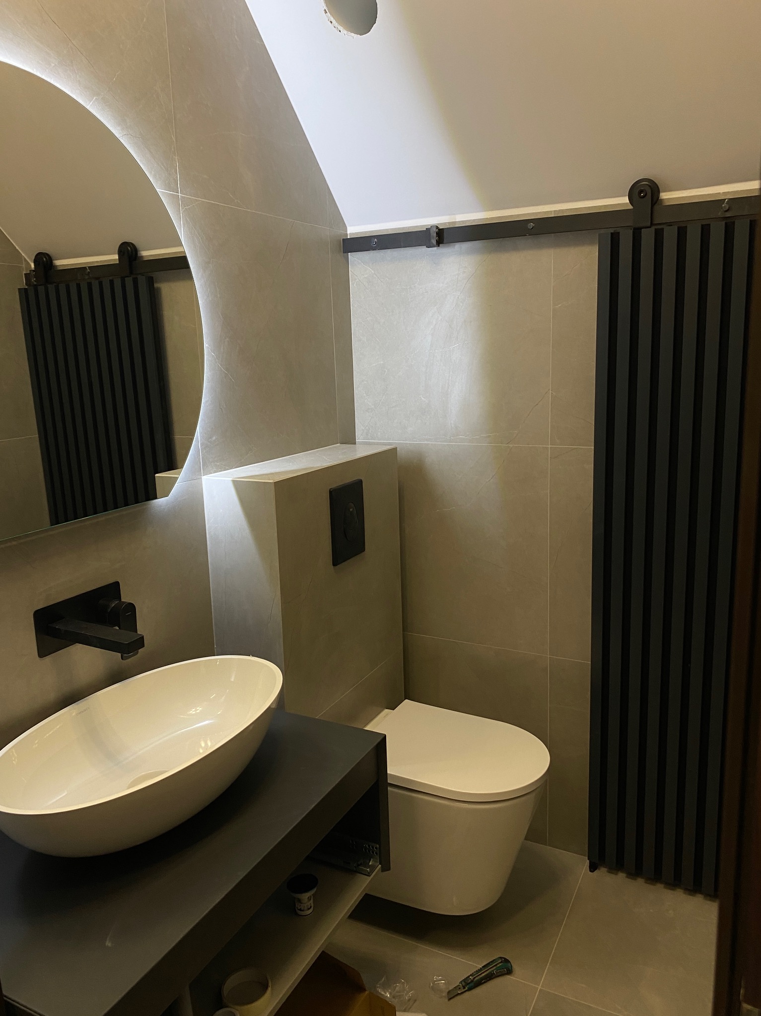 Nowoczesna łazienka na poddaszu z owalną umywalką nablatową, wiszącą toaletą i przesuwnymi panelami dekoracyjnymi w kolorze grafitowym, oświetlona okrągłym lustrem z podświetleniem.