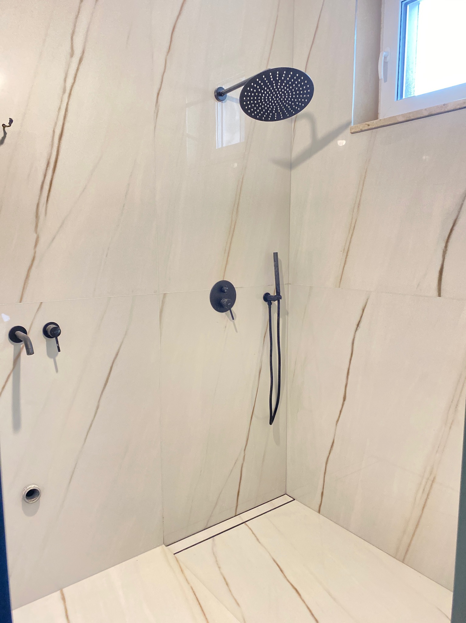 Kabina prysznicowa z białymi płytkami imitującymi marmur z brązowymi żyłkami, czarna deszczownica i bateria, odpływ liniowy przy ścianie, widoczne okno.