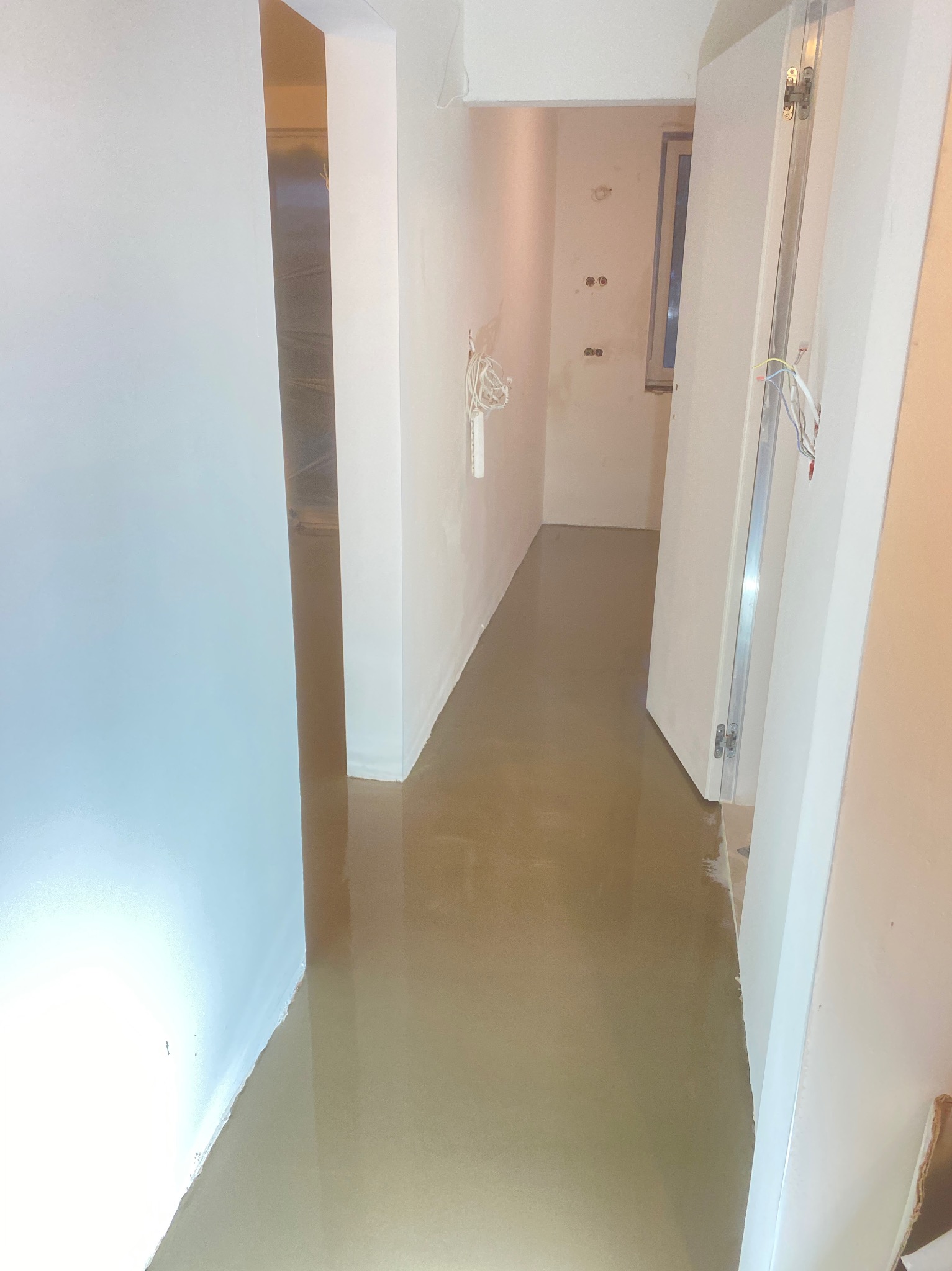 Korytarz w trakcie prac wykończeniowych: świeżo wylana, samopoziomująca wylewka podłogowa, ściany przygotowane do malowania, widoczne wystające kable elektryczne.