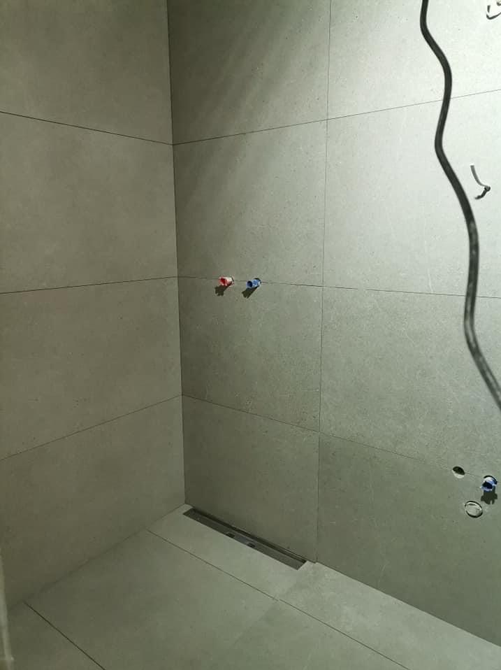 Szara łazienka w trakcie wykańczania, widoczne płytki wielkoformatowe, odpływ liniowy, wystające kable i rury z oznaczeniem ciepłej i zimnej wody.