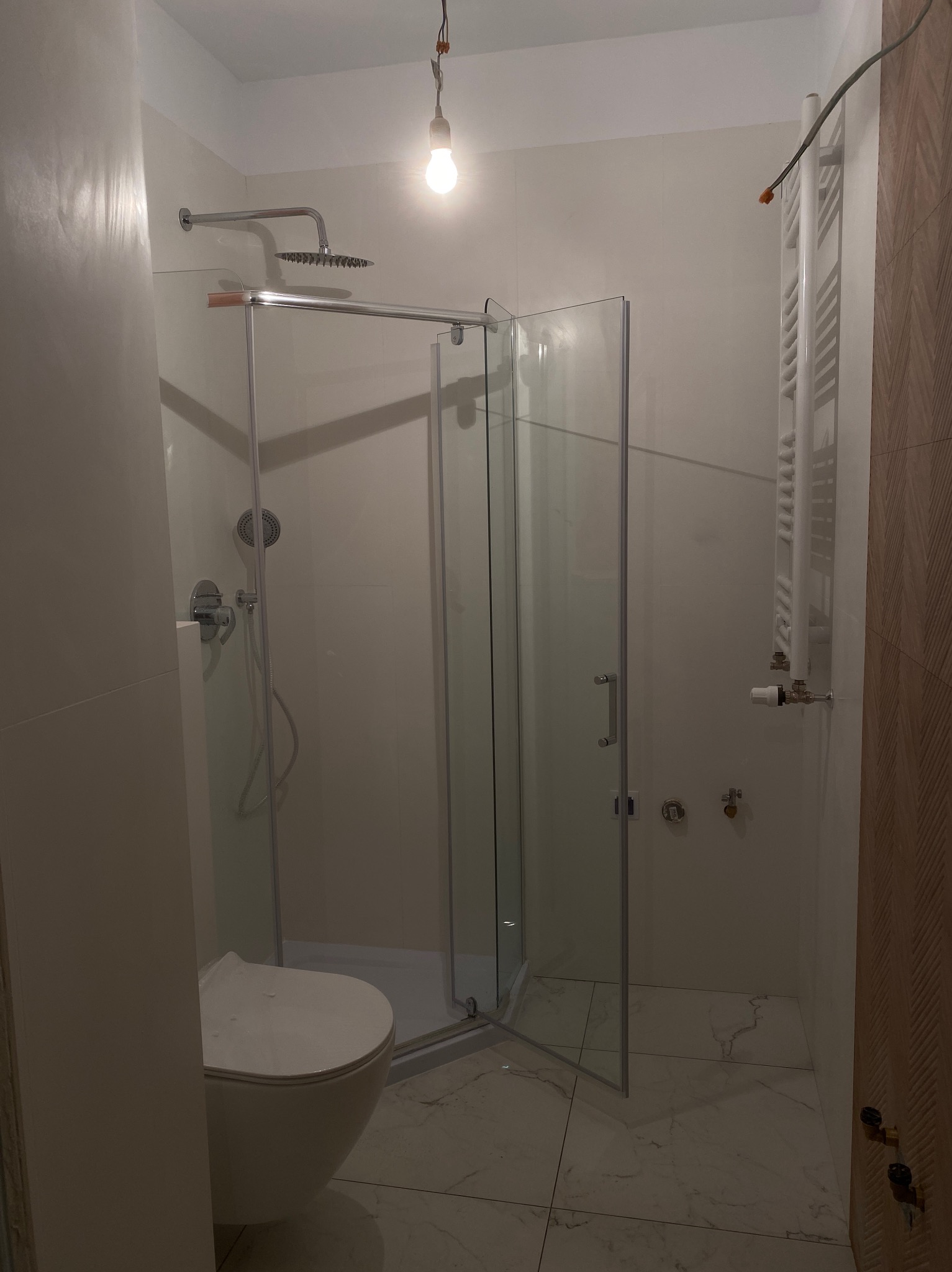 Nowoczesna łazienka z kabiną prysznicową typu walk-in, wiszącą toaletą i grzejnikiem drabinkowym, oświetlona wiszącą żarówką, marmurowe płytki podłogowe.