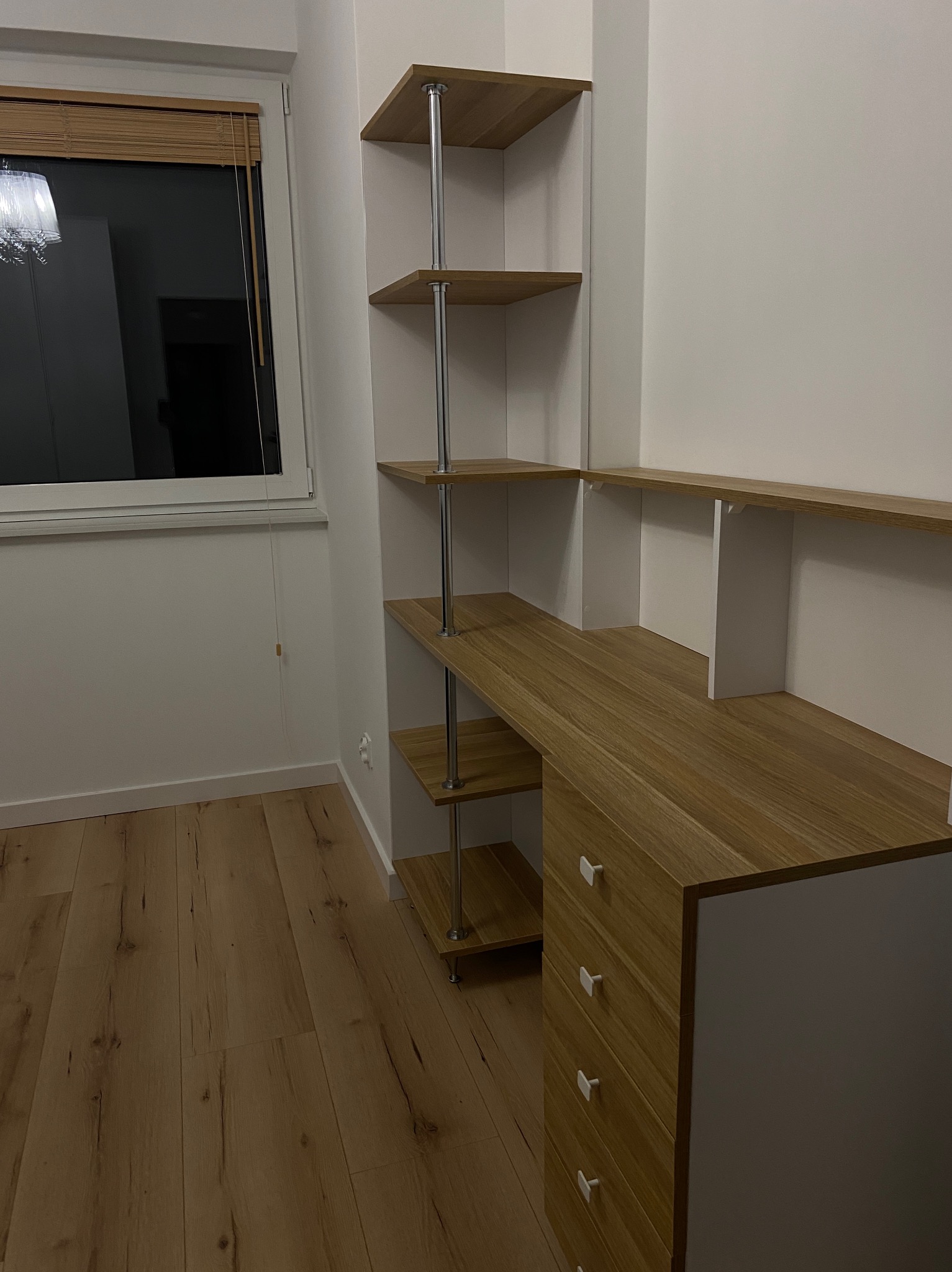 Nowoczesny pokój z drewnianą podłogą, biurkiem z szufladami i półkami na metalowych wspornikach, minimalistyczny styl, roleta w oknie.