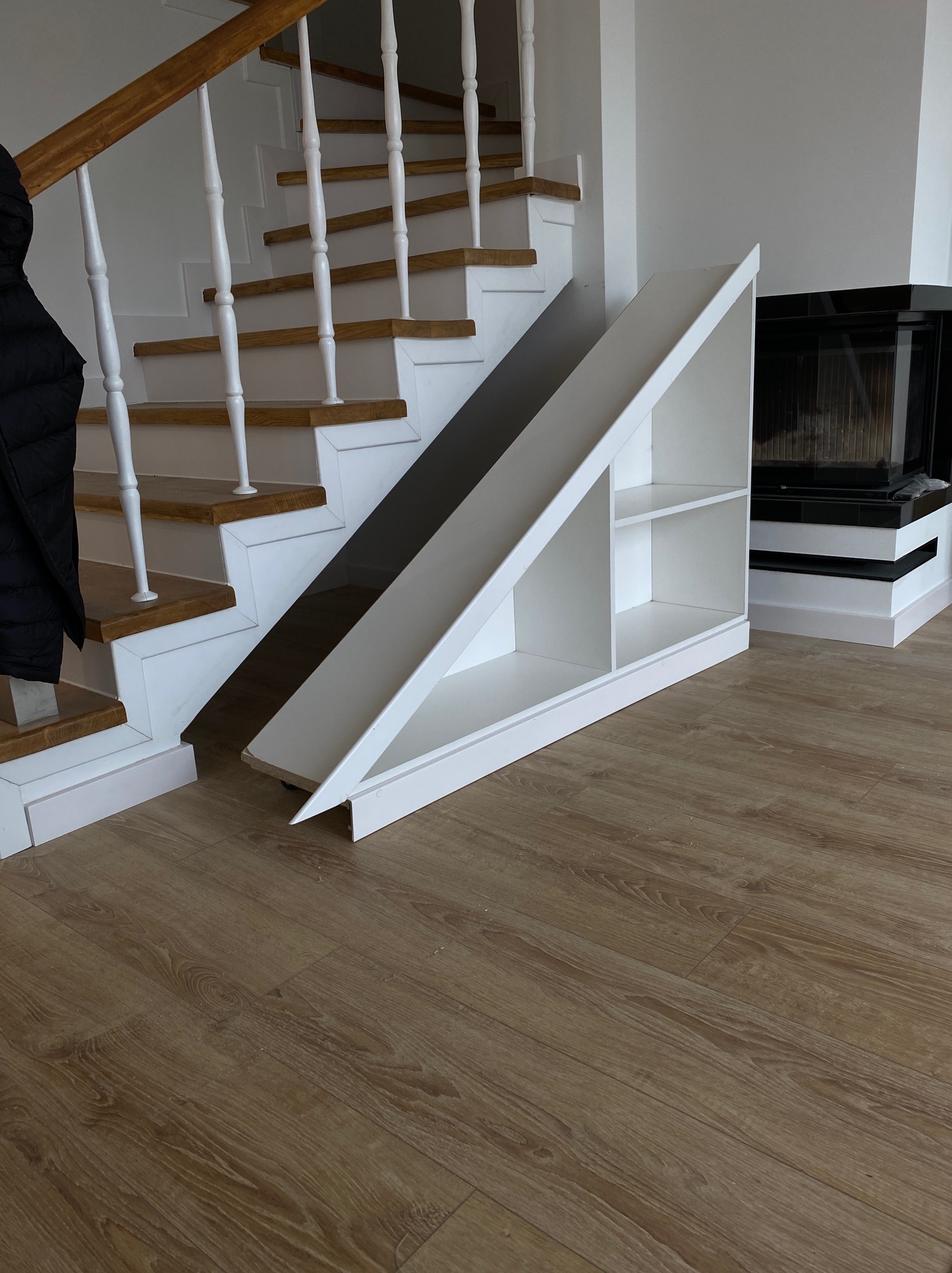 Białe schody z drewnianymi stopniami i balustradą, zintegrowane z trójkątnym regałem pod schodami, obok kominka w nowoczesnym wnętrzu.