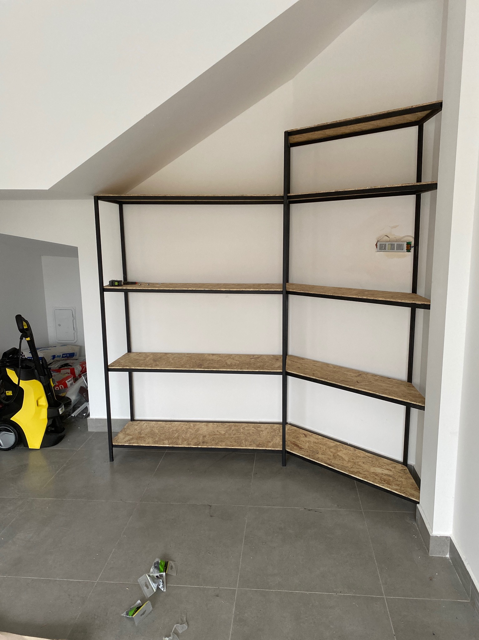 Metalowy regał z płytami OSB, dopasowany do skosu poddasza, widoczny w trakcie montażu, z niebieską taśmą mierniczą na jednej z półek i elementami montażowymi na podłodze.