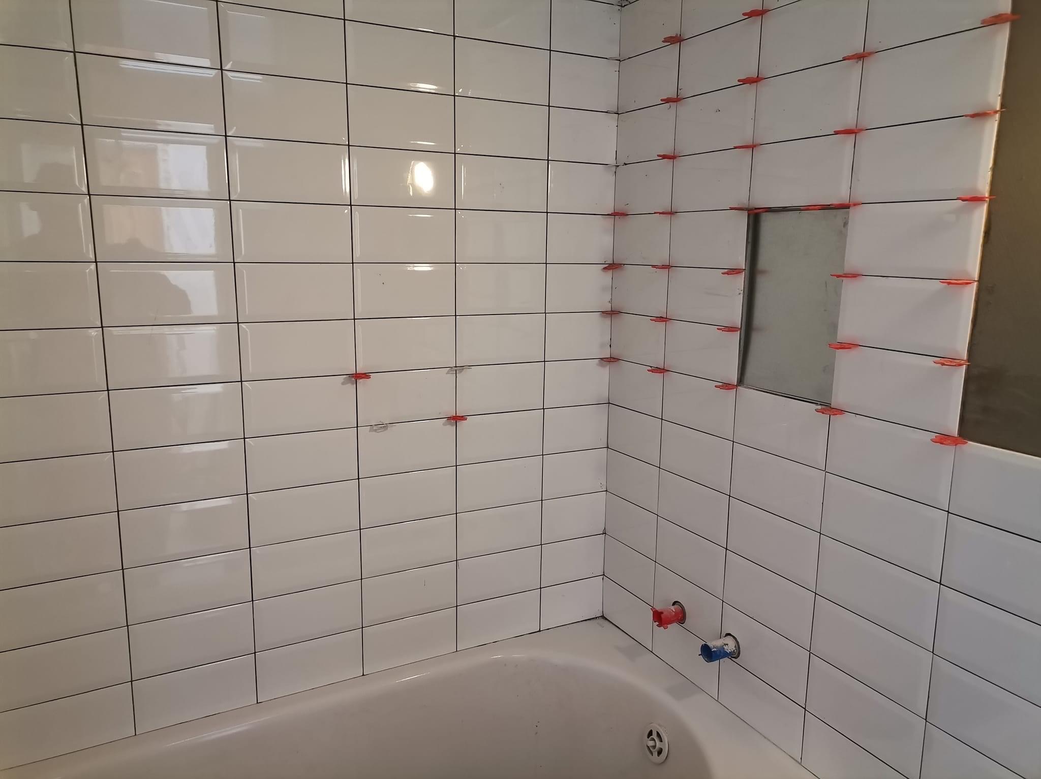 Białe, prostokątne płytki ścienne w trakcie układania w łazience, z widocznymi czerwonymi klinami poziomującymi, otwór na półkę bez płytek, oraz fragment wanny.