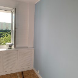 Mateusz Nowak - Świeżo pomalowana na jasnoniebiesko ściana w pokoju z parkietem, puszka farby lateksowej na parapecie przy otwartym oknie z widokiem na drzewa i blok.