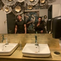 Mateusz Nowak - Łazienka z dwoma białymi umywalkami, złotym blatem i dekoracyjnym oświetleniem sufitowym. W lustrze odbija się mężczyzna robiący zdjęcie.