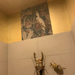 Mateusz Nowak - Fragment wnętrza z dekoracyjną tapetą w tropikalne wzory, złotą lamperią, dwoma metalowymi dekoracjami ściennymi w kształcie chrząszczy i okrągłym plafonem sufitowym.