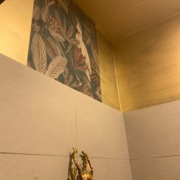 Mateusz Nowak - Fragment wnętrza z wzorzystą tapetą w tropikalne liście i kakadu, złotym wykończeniem sufitu i ścian oraz dekoracyjną figurką złotego chrząszcza.