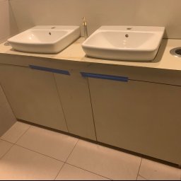 Mateusz Nowak - Dwie białe, prostokątne umywalki nablatowe na beżowej szafce z dwiema parami drzwi, zabezpieczonych niebieską taśmą malarską, obok dozownik mydła i otwór na kosz w blacie, ułożone na jasnych...