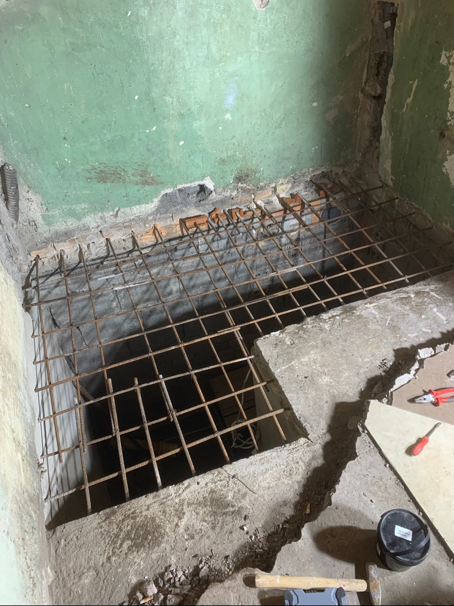 Przygotowanie podłoża: siatka zbrojeniowa ułożona w wykopie w pomieszczeniu z zieloną ścianą, widoczne narzędzia i wiadro z zaprawą.