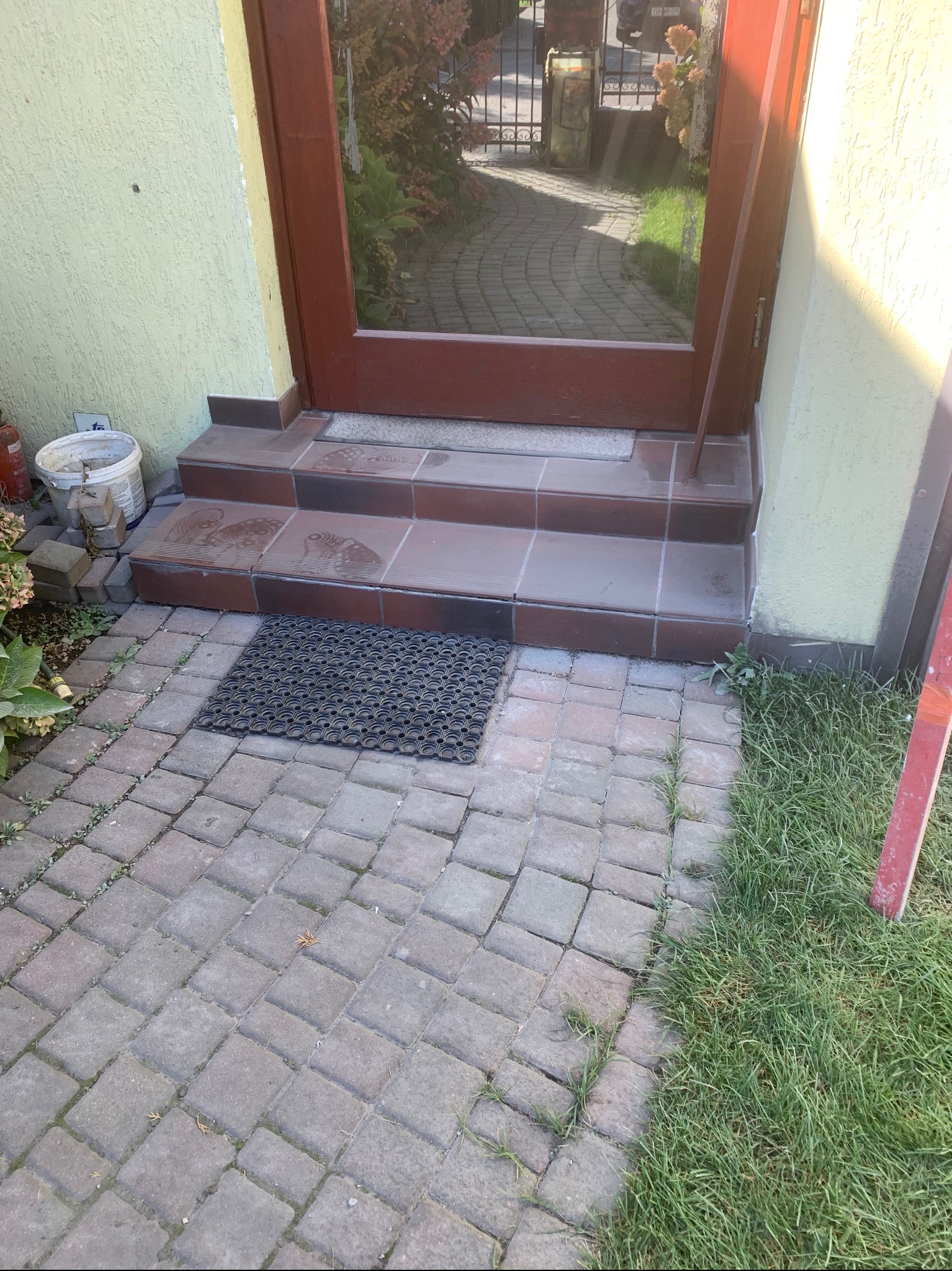 Wejście do domu z trzema stopniami wyłożonymi brązowymi płytkami z wzorem, czarna wycieraczka przed drzwiami, kostka brukowa i zielona trawa obok.