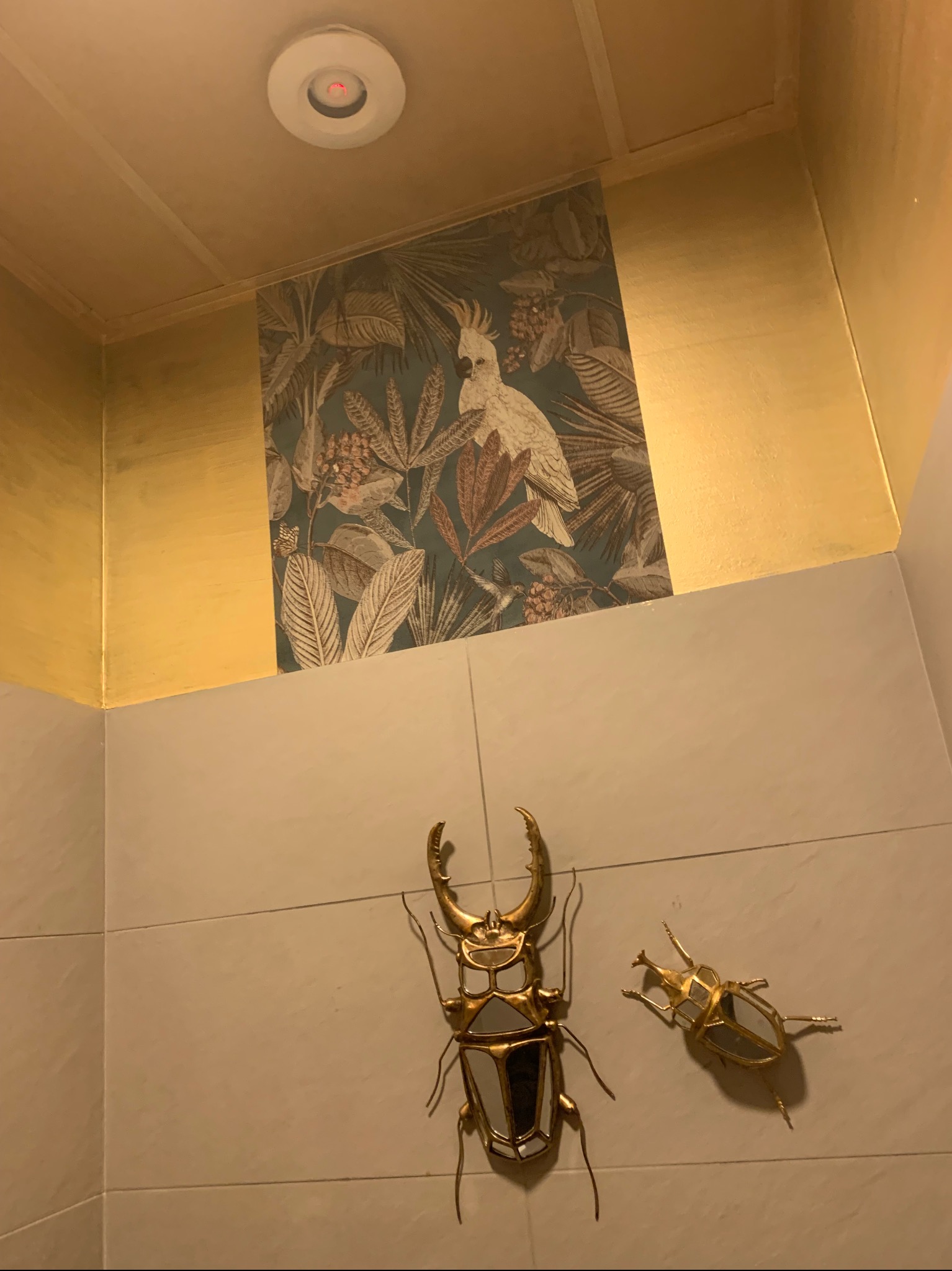 Fragment wnętrza z dekoracyjną tapetą w tropikalne wzory, złotą lamperią, dwoma metalowymi dekoracjami ściennymi w kształcie chrząszczy i okrągłym plafonem sufitowym.