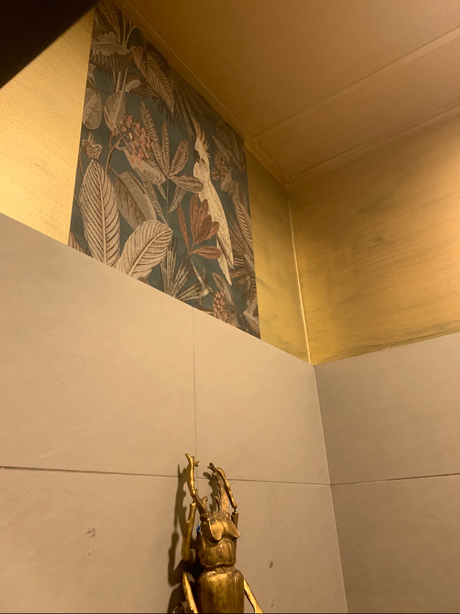 Fragment wnętrza z wzorzystą tapetą w tropikalne liście i kakadu, złotym wykończeniem sufitu i ścian oraz dekoracyjną figurką złotego chrząszcza.