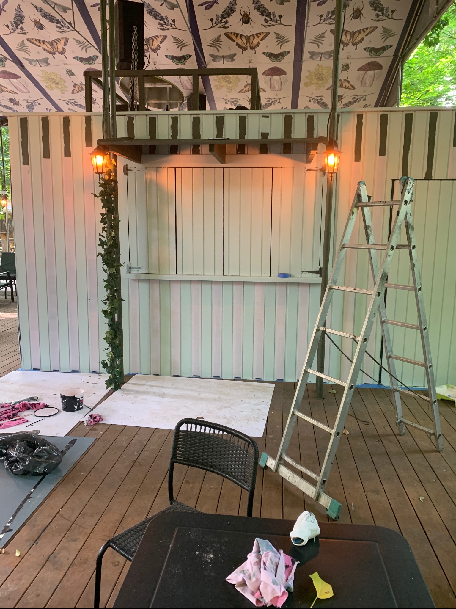 Malowanie ścian w paski na zewnątrz z użyciem drabiny, widoczne narzędzia malarskie, dekoracyjna lampa z bluszczem i sufit pokryty tapetą z motywem owadów i roślin.