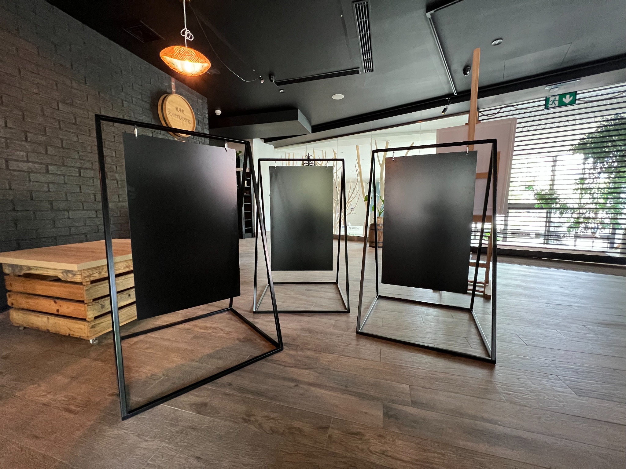 Trzy metalowe ramy z czarnymi panelami w nowoczesnym wnętrzu, widoczne drewniane elementy dekoracyjne i oświetlenie.