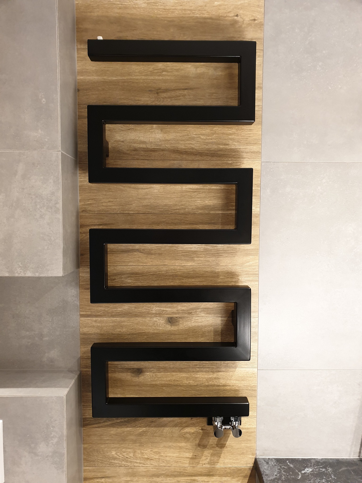 Czarny, dekoracyjny grzejnik łazienkowy w kształcie meandru, zamontowany na panelu imitującym drewno, obok szarych płytek.