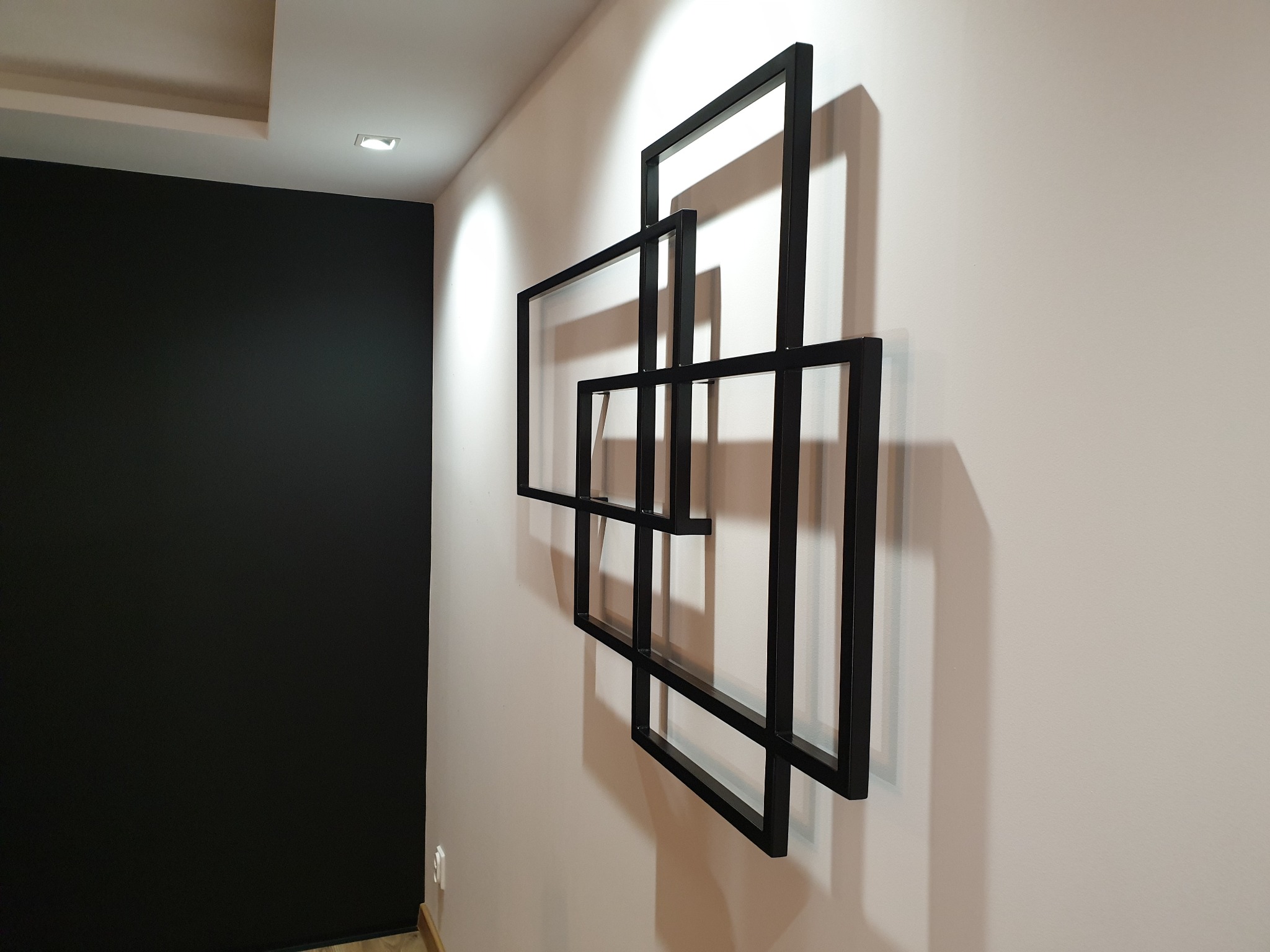 Metalowa dekoracja ścienna z czarnych, spawanych kwadratów o różnej wielkości, tworzących geometryczną kompozycję na tle białej ściany.